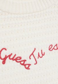 Kreemist kootud sviiter, millel on tekstureeritud kudumine ja sellele on alumises vasakus nurgas tikitud punane fraas "Guess Tu es". Ümmargune ribakrae.