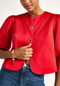 Rød bluse med korte ærmer og rund halsudskæring, dekoreret med sorte knapdetaljer, parret med blå denim jeans og et guldarmbånd om håndleddet.