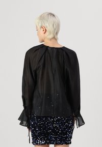 Svart gjennomsiktig bluse med lange ermer, strammende detaljer bak og bånd ved ermene, kombinert med et paljettbesatt skjørt i blått og svart.