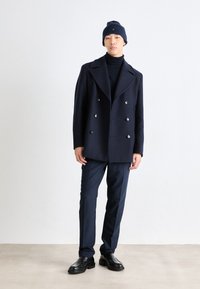 Manteau en laine bleu marine à double boutonnage avec des boutons en argent, associé à un col roulé noir, un pantalon bleu marine et des chaussures en cuir noires. Bonnet de la même couleur.