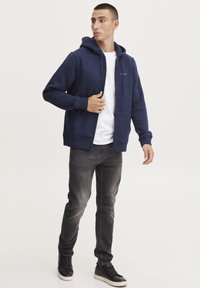 Jūras zilas krāsas zip-up kapuce ar priekšējām kabatām, ar regulējamu kapuci, uzvilkta virs baltā T-krekla, savienota ar pelēkajiem džinsiem un melnajiem kedām.