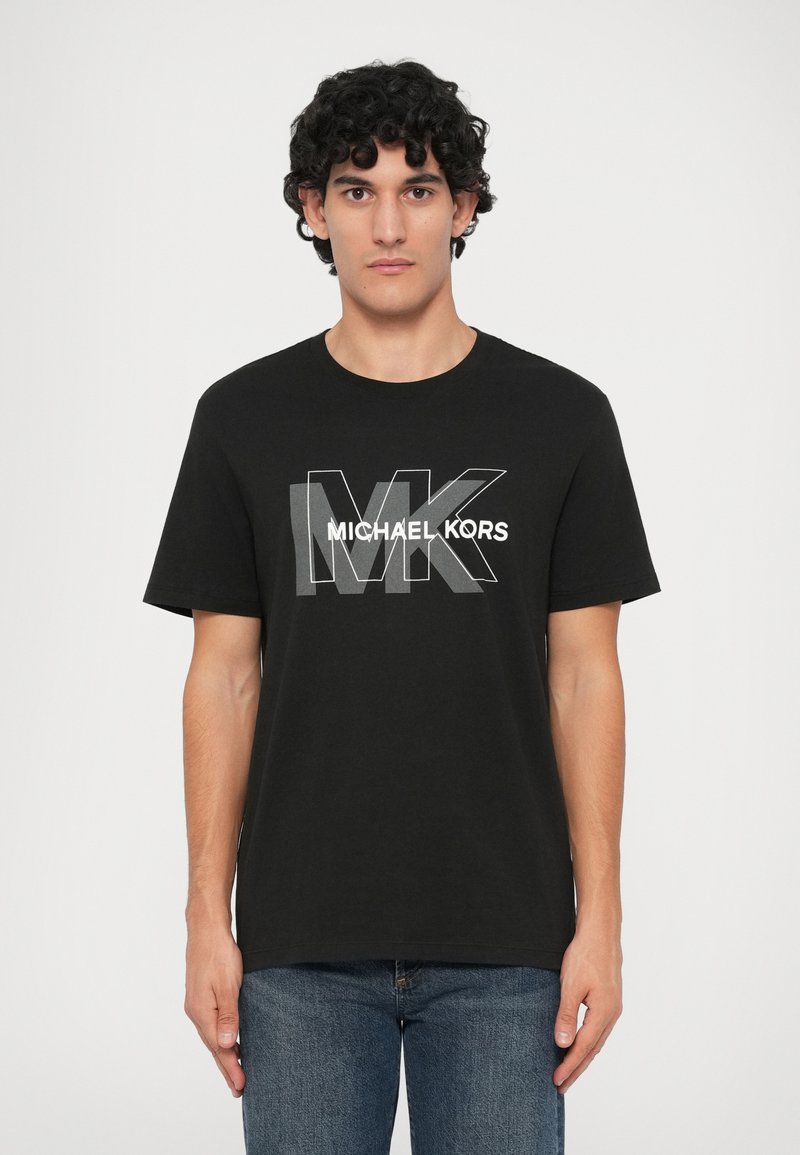 Muž s kučeravými čiernymi vlasmi, oblečený v čiernom tričku Michael Kors a modrých džínsach, stojí pred jednoduchým bielym pozadím.