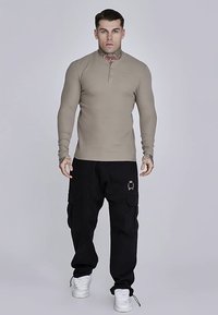 Bež dugi rukav s henley ovratnikom i uskim krojem, uparen s crnim cargo hlačama s džepovima i zlatnim logotipom.