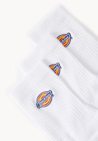 Három pár fehér bordázott zokni, amelyek kör alakú logóval rendelkeznek piros, sárga és kék díszítéssel, bemutatva a "Dickies" márkanevet.