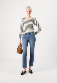 Top de manga larga a rayas, en beige claro y negro. Jeans de mezclilla azules, de corte recto. Bolsa de mano tejida marrón. Zapatos planos negros con detalles. Fondo blanco.
