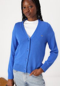 Blue cardigan featuring a v-neck, five buttons, ribbed hem and cuffs, made of lightweight knit material, worn over a white top.