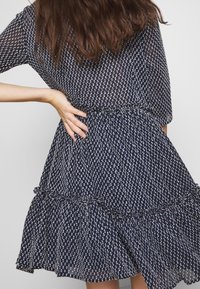 Mörkblå klänning med vitt prickmönster, korta ärmar, åtsittande midja och vågig volang nederkant. Tillverkad av lätt, semi-genomskinligt tyg.