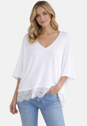 Femme portant un haut blanc ample à col en V avec manches larges et bordure en dentelle, associée à un jean bleu clair, debout devant un fond uni.