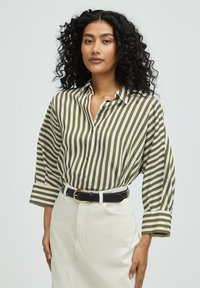 Striped shirt in olive green and white, features a loose fit, button closure, and rolled-up sleeves; paired with a black belt and white skirt.