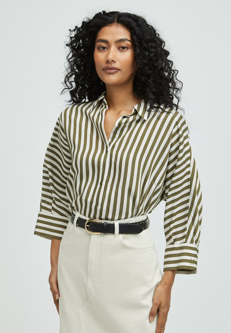 Striped shirt in olive green and white, features a loose fit, button closure, and rolled-up sleeves; paired with a black belt and white skirt.