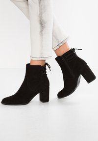 Persona che indossa stivaletti neri in suede con tacco a blocco e lacci frontali, abbinati a jeans chiari arrotolati.