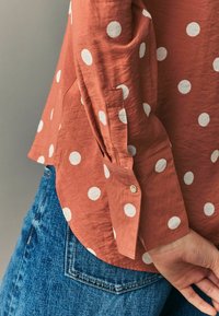 Rostfarbendes Hemd mit weißen Polka-Dots; lockere Passform, Knopfmanschetten-Detail, weicher, strukturierter Stoff, kombiniert mit blauen Jeans.