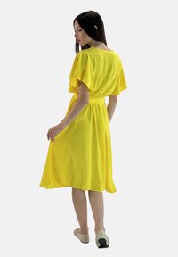 Gelbes knielanges Kleid mit kurzen, flatternden Ärmeln und tailliertem Schnitt. Leichter Stoff; fließende Textur und sanfter Fall.