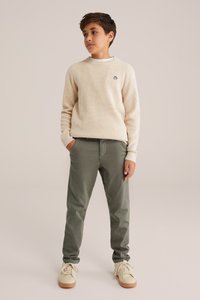 Bežs teksturēts džemperis ar mazu melnu emblēmu, apvienots ar zaļ�ām chinos biksēm un bežām kedām ar gumijas zoles, stāvot pret neitrālu fonu.