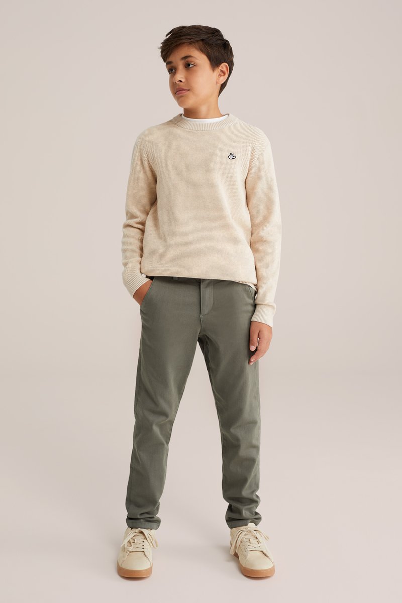 Bežs teksturēts džemperis ar mazu melnu emblēmu, apvienots ar zaļām chinos biksēm un bežām kedām ar gumijas zoles, stāvot pret neitrālu fonu.