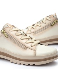 Baskets basses beige décontractées avec des accents beige clair, des lacets crème et des détails zippés latéraux sur une semelle en caoutchouc texturée.