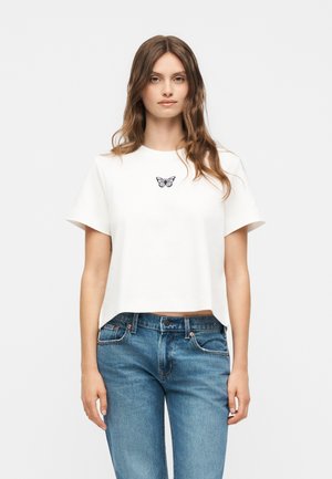 Ung kvinde med langt brunt hår iført en hvid cropped t-shirt med et lille sort sommerfugledesign og blå jeans, står oprejst.