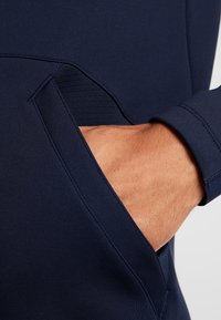Mørkeblå sweatshirt med et struktureret panel nær lommen. Materialet er glat; har tætsiddende manchetter og en sidelomme til hånden.