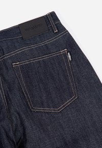 Jeans in denim scuro con etichetta in pelle sulla vita, cuciture arancioni a contrasto e una tasca posteriore con un piccolo marchio.