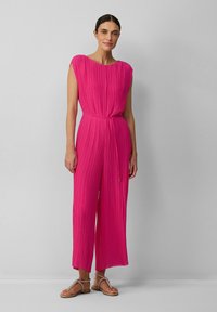 Fuchsia-gefüllter Jumpsuit mit rundem Ausschnitt, kurzen Ärmeln, einem gebundenen Bund und weitem Bein, kombiniert mit flachen Sandalen.