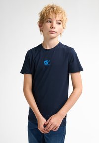 Námořnická modrá bavlněná tričko s kulatým výstřihem, krátkými rukávy a malým modrým logem na hrudi. Textura vypadá hladce a lehce.