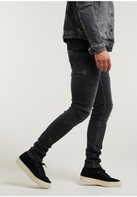 Černé slim-fit džíny s odřenými akcenty, spárované s černými semišovými teniskami se krémovou gumovou podrážkou. Džíny mají vybledlou texturu.