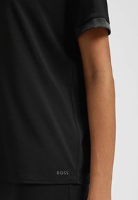 Zwart kortemouwenshirt gedragen door een persoon, met een close-up van de mouw en de zijzoom met een klein "BOSS" logo nabij de onderkant.