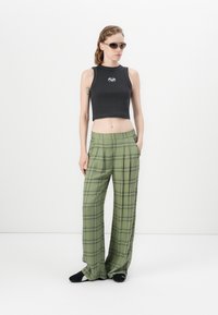 Woman wearing black sleeveless crop top, oversized green plaid pants, black shoes, and sunglasses standing against white background.