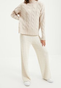 Personne portant un pull beige en maille torsadée et un pantalon large crema à texture, debout avec la main sur la hanche sur fond blanc.