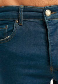 Pantalones vaqueros de mezclilla azul oscuro con un diseño ajustado, costuras visibles en un naranja contrastante y cierre delantero con botón.