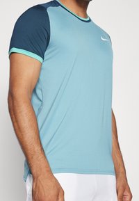 Camiseta deportiva de color azul claro con mangas marino. Presenta un cuello redondo, mangas cortas, una tela con textura y un pequeño logo blanco en el pecho.