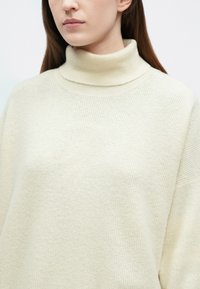 Kremiškai spalvos, pynuotas megztinis su aukštu kaklu, laisvo silueto ir minkštos tekstūros, turintis aukštą apykaklę ir be siūlių pečių dizainą.