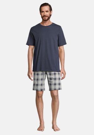 Man draagt een donkerblauw shirt met korte mouwen en grijs geruite knielange korte broek, staat blootsvoets tegen een effen lichte achtergrond.