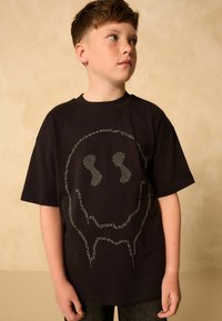 Tricou negru din bumbac, având un design mare cu o față zâmbitoare punctată pe față. Materialul pare moale și are o croială lejeră.
