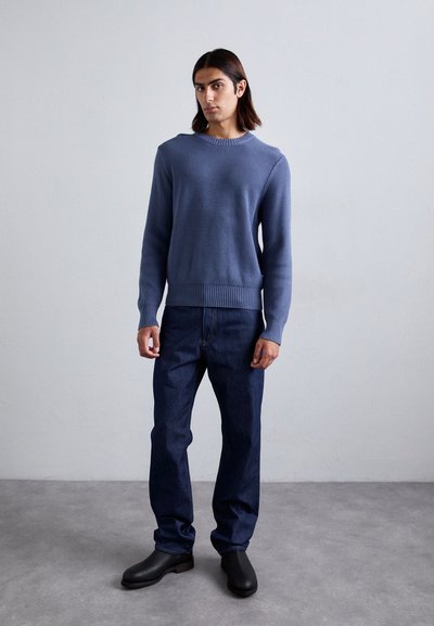 Suéter de punto azul con puños y dobladillo acanalados, combinado con jeans de mezclilla oscura y botines negros, de pie contra un fondo neutro.