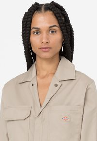 Camisa de trabajo beige con cuello puntiagudo, bolsillo en el pecho con un parche de logo y una textura de tela duradera. Botones en la parte delantera.