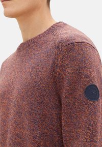 Pull en tricot texturé en fil multicolore avec des nuances de violet et d'orange ; doté d'un col rond et d'un patch en tissu sur la manche.