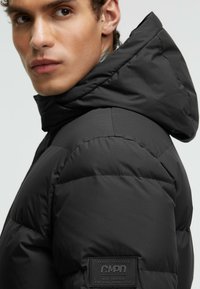 Black puffer jacket with a high collar and hood. Features quilted texture and a branded patch on the left side. Smooth synthetic material.