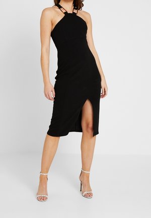 Schwarzes ärmelloses Kleid mit figurbetonter Silhouette, vorderem Schlitz und charakteristischen Neckholder-Trägern. Kombiniert mit silbernen Sandalen mit Absatz.