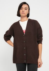 Καφέ ριγέ ζακέτα με V-λαιμόκοψη, φαρδιά εφαρμογή και μεγάλα σκούρα κουμπιά. Μακριά μανίκια με χαλαρή μανσέτα.