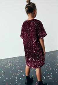 Bordeaux farvet sequin kjole med afslappet pasform, korte ærmer og nøglehul detalje bagpå, parret med sorte ankelstøvler på en farverig konfetti gulv.