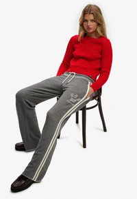 Rød kabelstrikket sweater med lange ærmer, kombineret med grå joggingbukser med hvide sidelommer og et grafisk tryk. Brune sko medfølger.