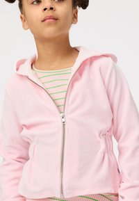 Sweatshirt rosa claro com fecho, em tecido felpudo, apresentando capuz com cordão e laterais franzidas, usado sobre uma camisa listrada em verde e branco.