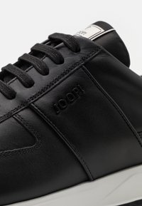 Svarta lädersneakers med platta snören, präglad logotyp på sidan och en vit gummisula. Slät textur med sydda detaljer.