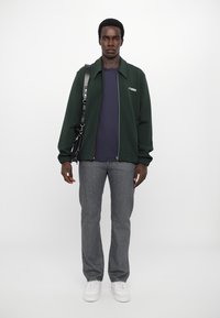Mørkegrønn lett jakke med glidelås, marineblå skjorte under, grå jeans og hvite sneakers. Inkluderer en svart skulderveske.