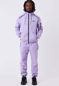 Ensemble de sport violet comprenant une veste zippée avec des accents noirs et des poches latérales, assortie à un pantalon avec des poignets élastiques.