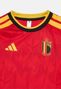 Κόκκινη φανέλα ποδοσφαίρου με κίτρινες λεπτομέρειες, που διαθέτει μαύρο �γιακά και τρεις μαύρες ρίγες. Περιλαμβάνει το βέλγικο έμβλημα και το λογότυπο της Adidas.