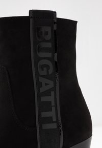 Botín negro hasta el tobillo con parte superior de ante, detalles negros brillantes y un prominente logo "BUGATTI" en la lengüeta. Diseño de punta redondeada.