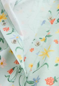 Gros plan sur un tissu vert clair avec des motifs floraux jaunes, orange et bleus et des boutons-pression argentés sur une ouverture de vêtement.