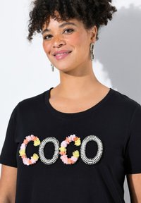 Camiseta de algodón negra con el texto "COCO" adornado con apliques florales coloridos y strass. Cuello redondo, mangas cortas.
